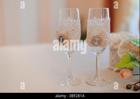 Nahaufnahme zweier Sektflöten, die für eine Hochzeit dekoriert sind, mit komplizierten Spitzendetails, weichen pfirsichfarbenen Bändern, die mit Vintage-Broschen verbunden sind, und sanften Details Stockfoto