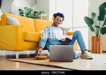 Junger Mann genießt Freizeit, Musik zu Hause zu hören, während er liest und sich in einem stilvollen Wohnzimmer entspannt Stockfoto