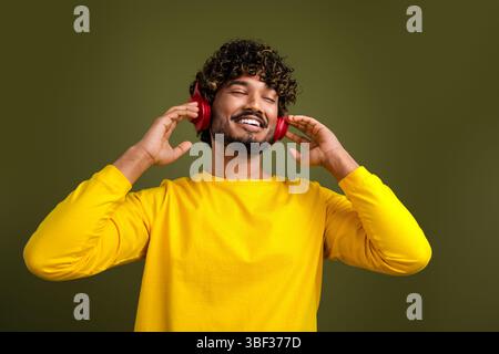 Junger Mann, der Musik mit roten Kopfhörern genießt, trägt ein gelbes Hemd vor grünem Hintergrund Stockfoto