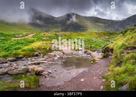 Eine ruhige und ruhige Bergszene von Carrauntoohil entfaltet sich mit einem sanft fließenden Bach, umgeben von üppigem, lebhaftem Grün und dramatischen Wolken Stockfoto