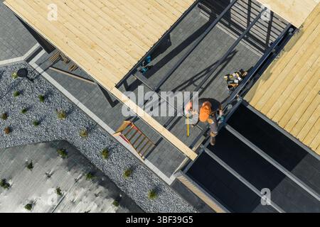 Zwei Arbeiter installieren sorgfältig Holzbalken auf einem Gebäudedach und zeigen Teamwork und Präzision auf einer belebten Baustelle unter klarem Himmel. Stockfoto