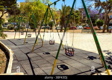 Eine Reihe leerer Kleinkinder schwingt an einem sonnigen Tag auf einem ruhigen Spielplatz in einem Stadtpark. Schatten fallen auf die Gummimatte, mit Bäumen, Autos und Gebäuden Stockfoto
