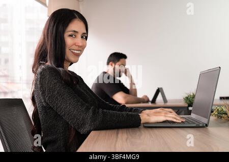 Selbstbewusste Frau lächelt, während sie ihr Kinn auf der Hand hält, während sie am Laptop arbeitet. Ein durchdachter und dennoch optimistischer Moment in einem modernen Arbeitsbereich. Stockfoto