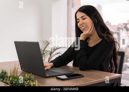 Frau lächelt, während sie an einem Laptop in einem gemeinsamen Büro arbeitet. Ruhige, fokussierte Umgebung mit modernem Stil und Tageslicht. Professionell und ruhig. Stockfoto