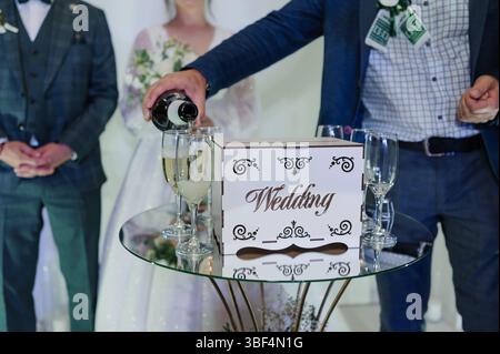 Ein Bräutigam in einem stilvollen Anzug gießt Champagner in Gläser, die auf einem silbernen Tablett gehalten werden, während die Braut in einem weißen Kleid lächelt, eine Feier mit einem Dekor Stockfoto