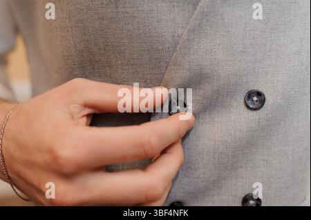 Eine Nahaufnahme zeigt einen Mann, der elegant die Knöpfe einer grauen Tweed-Weste anschließt und das strukturierte Material und die raffinierten Details des Su betont Stockfoto