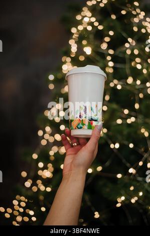 Eine hellhäutige Hand hält eine weiße Papierkaffeetasse sanft in die Wiege, die mit einem lebhaften geometrischen Weihnachtsmuster verziert ist, während sie vom sanften Leuchten beleuchtet wird Stockfoto