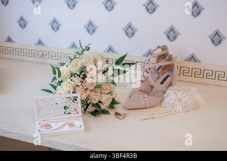 Eine wunderschöne Aufnahme zeigt Brautschuhe, einen frischen Blumenstrauß, Eheringe und zarten Schmuck auf einem hellen, gemusterten Sideboard, c Stockfoto