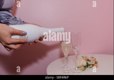 Eine stilvolle Frau schenkt Champagner in zwei Gläser vor einem sanften rosa Hintergrund, ergänzt durch zarte Rosen, die für eine feierliche Stimmung perfekt sind Stockfoto