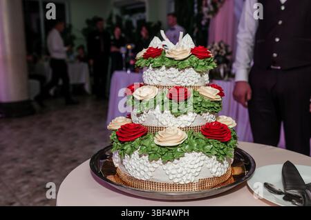 Eine wunderschön dekorierte, dreistöckige weiße Hochzeitstorte mit aufwändigen Zuckerguss und leuchtenden roten Rosenverzierungen, zentriert an einem Abend im Freien Stockfoto