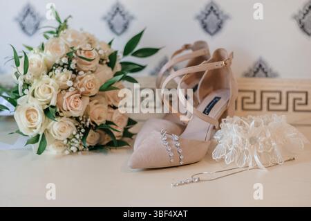 Eine Nahaufnahme eleganter beiger Hochzeitsschuhe, ein weicher pfirsichfarbener Blumenstrauß und zarten Schmuck auf einem hellen Holztisch mit gemustertem Muster Stockfoto
