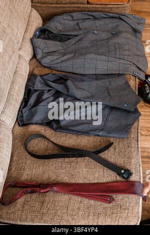 Eine graue karierte Anzugsjacke, Hose und Weste ist lässig über eine braune Couch mit einem braunen Ledergürtel und Hosenträgern gelegt, was eine entspannte Mode andeutet Stockfoto