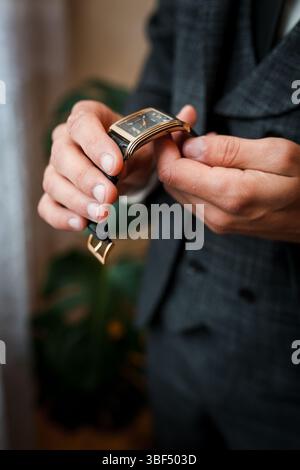 Eine Nahaufnahme zeigt einen gut gekleideten Mann, der eine elegante rechteckige Golduhr mit einem schwarzen Lederarmband befestigt und einen formellen grauen Tweed zeigt Stockfoto