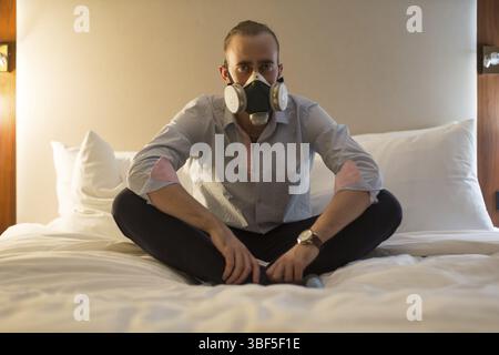 Hübscher junger, stilvoller kaukasischer Mann in Atemschutzmaske, der auf dem Bett im Schlafzimmer sitzt Stockfoto