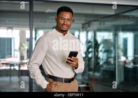 Glücklicher professioneller afroamerikanischer Geschäftsmann, der im Büro mit dem Telefon steht. Stockfoto