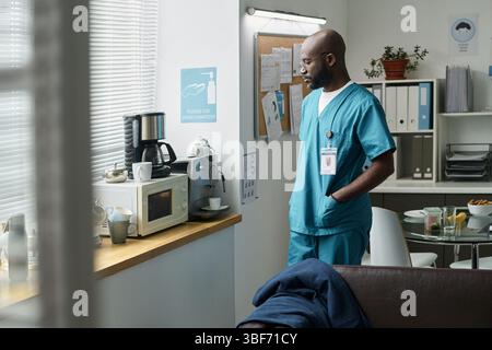 Afroamerikanische männliche Krankenschwester steht im Pausenraum des Krankenhauses neben Kaffeemaschine und Anschlagtafel. Blaue Peelings tragen und Einwegkaffeetasse halten, während der Pause entspannen Stockfoto