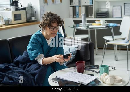 Porträt eines Arztes, der auf der Couch im Büro sitzt, auf das Telefon blickt und von Dokumenten, Kopfhörern und Kaffeetassen umgeben ist, möglicherweise nach einer langen Schicht Stockfoto