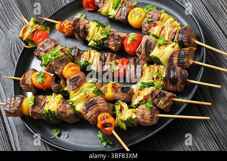 Spieße von gegrilltem Rindfleisch mit Zucchini-Bändern und Tomaten, serviert auf Teller über dunklem Holz, Foto von echtem Essen, Stockfoto