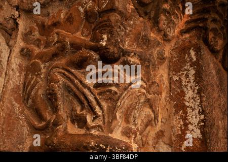 Ein Mann sitzt mit einem langen Gegenstand - vielleicht einem Knochen - zwischen den Zähnen auf einer aus dem 12. Jahrhundert stammenden Hauptstadt, die Tympanon und Archivolte des romanischen Nordportals der spanischen Romanik von Iglesia de San Salvador oder der Kirche San Salvador in Agüero, Provinz Huesca, Aragon, Spanien, unterstützt. Reich geschnitzte Tür, die dem Bildhauer zugeschrieben wird, der als Meister von Agüero bekannt ist. Stockfoto