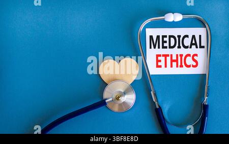 Medizinethik-Symbol. Konzeptworte medizinische Ethik auf der schönen weißen Karte. Holzherz und Stethoskop. Wunderschöner blauer Hintergrund. Medizinisch gesund Stockfoto
