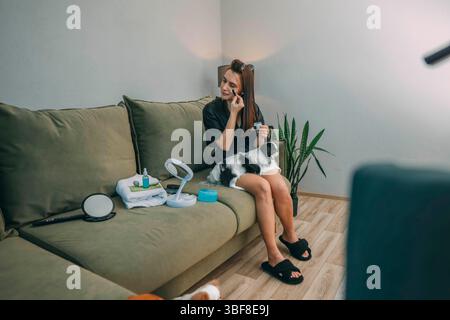 Frau, die Gesichtscreme unter den Augen mit einem japanischen Kinnhund auf dem Schoß aufträgt, in gemütlichem Wohnzimmer mit Pflanzen, Wellness-Lifestyle, Tierpflege, Schönheitsprodukten Stockfoto