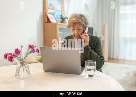 Eine ältere Frau, die zu Hause einen Laptop benutzt, während sie telefoniert. Technologiekonzept, Online-Shopping, digitale Kompetenz für Senioren und aktives Altern Stockfoto