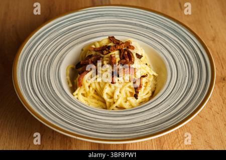 Eine wunderschön überzogene Portion Spaghetti Carbonara mit knusprigem Pancetta auf einer gestreiften Keramikplatte. Die cremige Soße und die goldenen Töne erzeugen einen Stockfoto