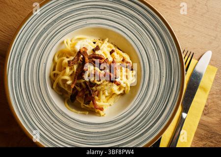 Eine wunderschön überzogene Portion Spaghetti Carbonara mit knusprigem Pancetta auf einer gestreiften Keramikplatte. Die cremige Soße und die goldenen Töne erzeugen einen Stockfoto