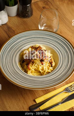 Eine wunderschön überzogene Portion Spaghetti Carbonara mit knusprigem Pancetta auf einer gestreiften Keramikplatte. Die cremige Soße und die goldenen Töne erzeugen einen Stockfoto