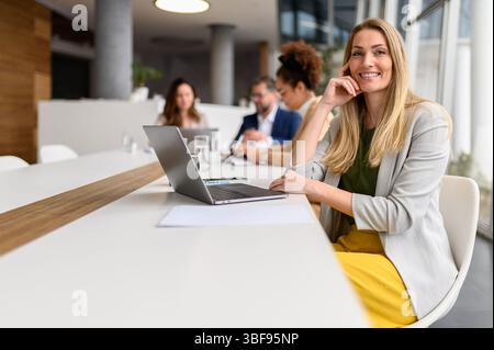 Porträt einer erfolgreichen Geschäftsfrau, die am Laptop arbeitet, während Geschäftsleute im Hintergrund zusammenarbeiten Stockfoto