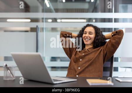 Eine entspannte Frau lächelt, lehnt sich zurück in ihrem Bürostuhl mit Händen hinter dem Kopf, in der Nähe eines Laptops auf einem Schreibtisch. Stockfoto