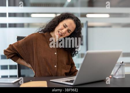 Eine Frau mit lockigen Haaren hat Rückenschmerzen, während sie an ihrem Laptop am Schreibtisch arbeitet. Sie ist sichtlich unwohl und hält sie zurück. Stockfoto