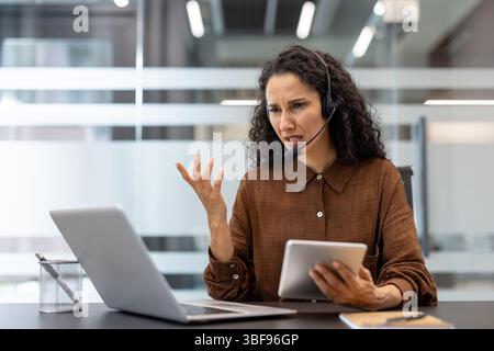 Ängstliche Frau, die ein Headset trägt, hat einen wütenden Ausdruck, während sie an einem Computer oder Tablet arbeitet. Sie ist frustriert. Stockfoto