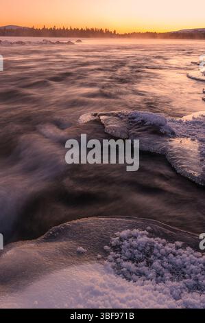 Der Fluss fließt unter Eis mit sichtbaren Formationen, während ein atemberaubender Sonnenuntergang den Himmel in warmen Farben färbt. Nebel steigt aus dem Wasser auf und verstärkt den Ser Stockfoto