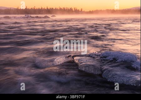Der Fluss fließt unter Eis mit sichtbaren Formationen, während ein atemberaubender Sonnenuntergang den Himmel in warmen Farben färbt. Nebel steigt aus dem Wasser auf und verstärkt den Ser Stockfoto
