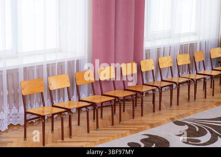Einfache Holzstühle stehen in einem hell beleuchteten Raum mit großen Fenstern. Weiche Vorhänge hängen neben polierten Holzböden und schaffen ein warmes und warmes Ambiente Stockfoto