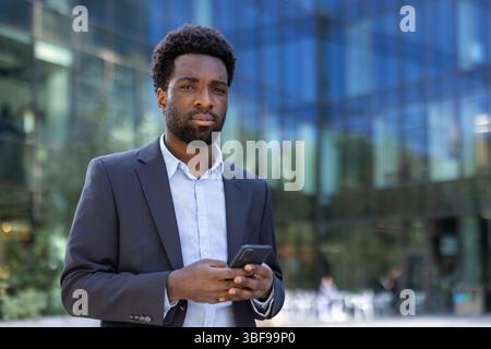Ein klug gekleideter afroamerikanischer Mann hält sein Smartphone und posiert selbstbewusst vor einem modernen Bürogebäude. Stockfoto
