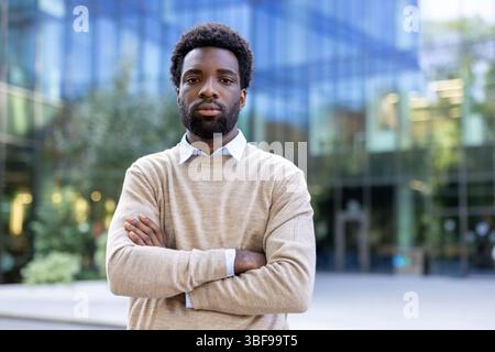 Ein afroamerikanischer Mann steht selbstbewusst mit gekreuzten Armen vor einem Bürogebäude. Er schaut mit ernstem Ausdruck in die Kamera. Stockfoto