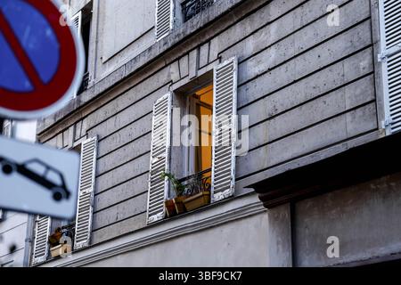 Nahaufnahme eines Pariser Apartmentfensters mit klassischen weißen Fensterläden, einem kleinen Balkongeländer mit Blumenkasten und einer warmen, einladenden Lichtbeleuchtung Stockfoto