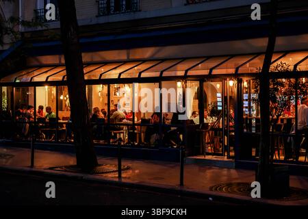 Blick auf das geschäftige Bistro in Paris bei Nacht. Die Glasfront zeigt ein warmes Interieur voller Menschen, die essen, trinken und sozial unterwegs sind Stockfoto