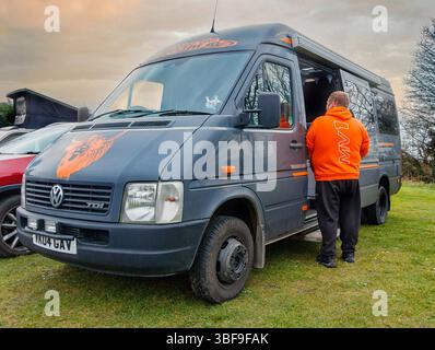 Ein Mann mit orangefarbener Jacke steht neben einem Volkswagen Crafter Van mit personalisierten Grafiken, die bei Sonnenuntergang auf dem Gras geparkt sind Stockfoto