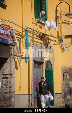 Eine typische neapolitanische Straßenszene. Ein Mann, der uns aus einem Fenster im Zentrum von Neapel blickt und Passanten auf der Straße unter den bunt bemalten Gebäuden auf dem Gerolomini-Platz in Neapel, Italien, spaziert. Stockfoto