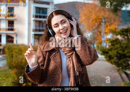 Porträt einer glücklichen jungen Kaukasierin im Mantel, die Musik mit Kopfhörern hört und im Park singt. Das Konzept von Glück und Freiheit. Stockfoto