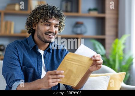 Ein fröhlicher Mann, der einen Umschlag öffnet, aufgeregt und glücklich wirkt. Er trägt ein Jeansshirt in einer bequemen Umgebung. Stockfoto