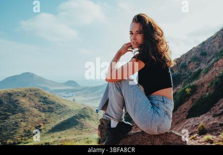 Junge schöne Frau, die auf Stein auf Berglandschaftshintergrund sitzt Stockfoto