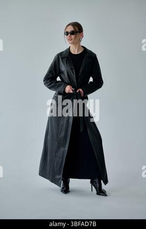 Modische junge Frau in schwarzem Kleid und schwarzem Ledermantel und Sonnenbrille, die im Studio posiert Stockfoto