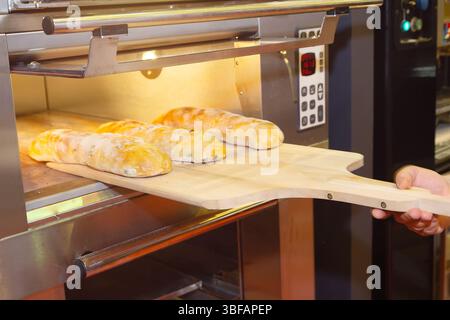 Der Bäcker nimmt aus dem Elektroofen heißes, frisch gebackenes Brot auf einem Holztablett heraus. Lebensmittelindustrie Stockfoto