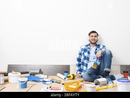 Gutaussehender bärtiger Mann, der zu Hause Musik in Kopfhörern hört. Hausumbau nach Umzugskonzept. Malwerkzeuge und Material auf dem Boden. Junger kauca Stockfoto