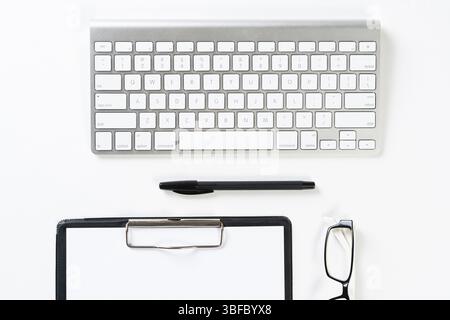 Draufsicht auf einen gut organisierten Arbeitsplatz mit Bürozubehör. Zwischenablage mit leerer Papierseite, Brille und drahtloser Tastatur auf weißer Oberfläche. Lehrerin Stockfoto