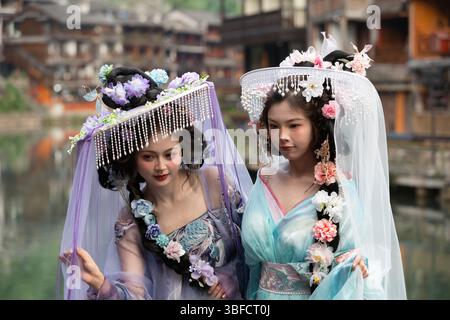 Feng Huang, China - 25. April 2025 : schöne chinesische Frauen in traditioneller Volkstracht, die Hände halten und für ein Bild vor Wald posieren Stockfoto